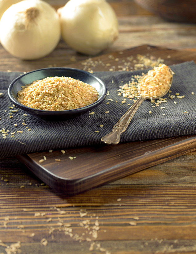 Dried minced onion flakes in a small black bowl and silver spoon next to whole white onions